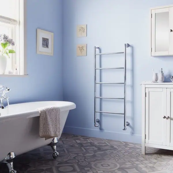 Chrome towel radiator in a modern bathroom with vintage bathtub and white cabinetry, light blue walls, and patterned floor tiles.
