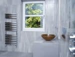 Sleek modern bathroom with gray tiled walls, wall-mounted towel radiator, round wooden sink, and a large window with a view of green foliage.