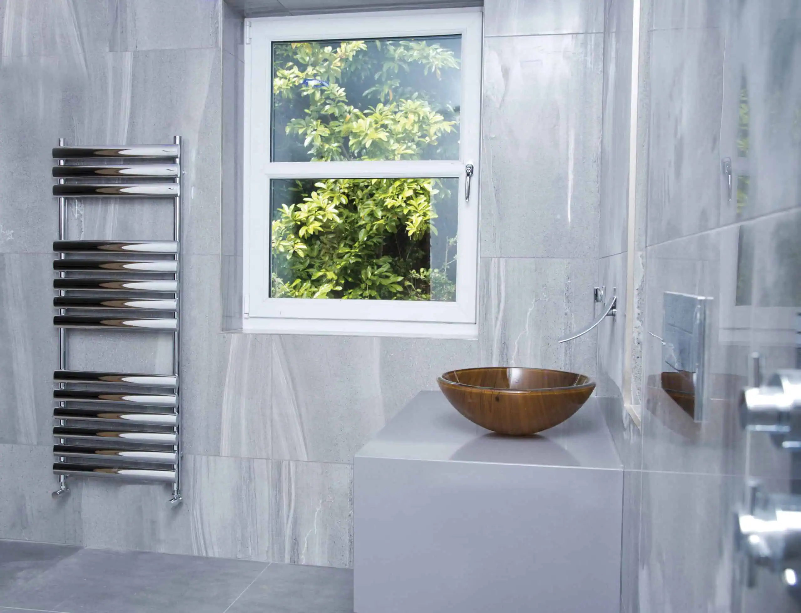 Dorney-Towel-Rail-Chrome-1200x500-Lifestyle-rgb-scaled-2.jpg Sleek modern bathroom with gray tiled walls, wall-mounted towel radiator, round wooden sink, and a large window with a view of green foliage.