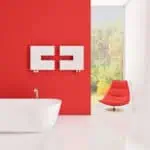 Modern bathroom interior with red accent wall, white bathtub, and stylish lounge chair, bright natural light.
