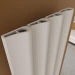 Elongated white radiator with curved panels and top openings, installed on a brown wall.