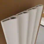 Elongated white radiator with curved panels and top openings, installed on a brown wall.